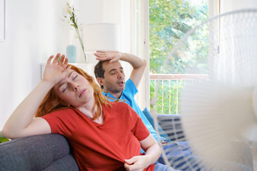 A man and woman sitting on the couch in a hot house with a fan blowing on them.