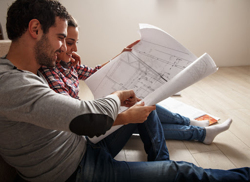 A man and woman sitting on the floor in a home and looking at blueprints for a house.