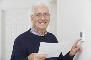 A smiling man holding a paper bill as he adjusts the thermostat in a home.