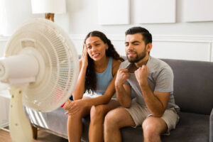 A man and woman sitting in front of a standing fan trying to cool off in a hot living room.