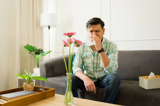 A man blowing his nose as he sits on a couch in front of a vase of flowers.